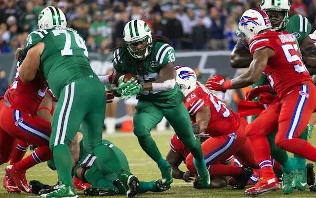 An American football play: one team in all-green uniforms carries the ball through a line of players in all-red uniforms.